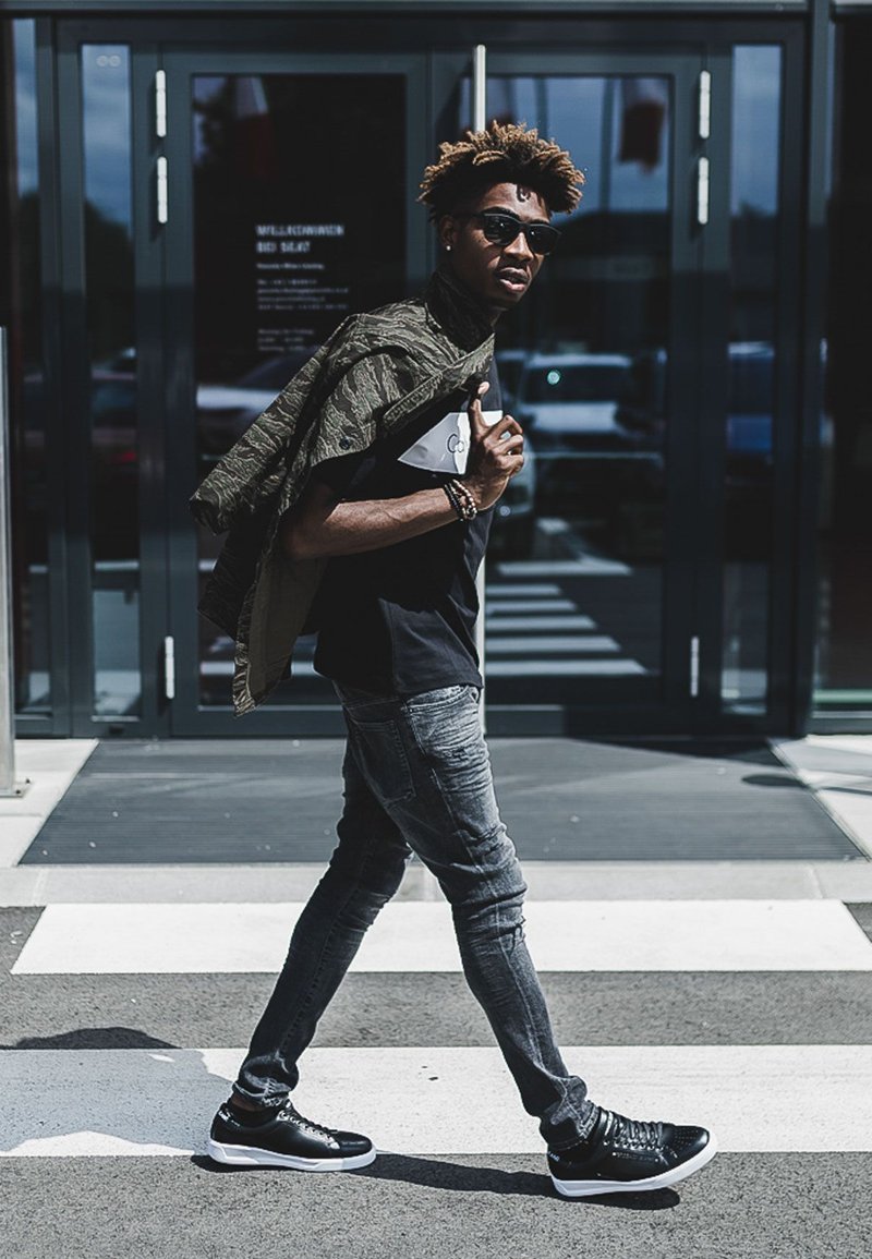 Black t-shirt with a white logo, grey slim-fit jeans, black trainers, and an olive green patterned jacket draped over the shoulder.