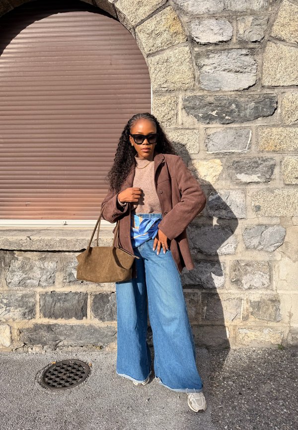 Donna con capelli ricci, occhiali da sole, giacca marrone, top beige, jeans larghi blu e sneakers bianche, in piedi contro un muro di pietra sotto la luce del sole.