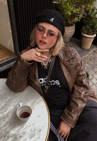 Barna bőrkabát, fekete beret, rózsaszín napszemüveg, fekete Adidas póló. Figyelemre méltó ékszerek charmokkal és mintás hosszú körmökkel. Márvány asztal csészével.