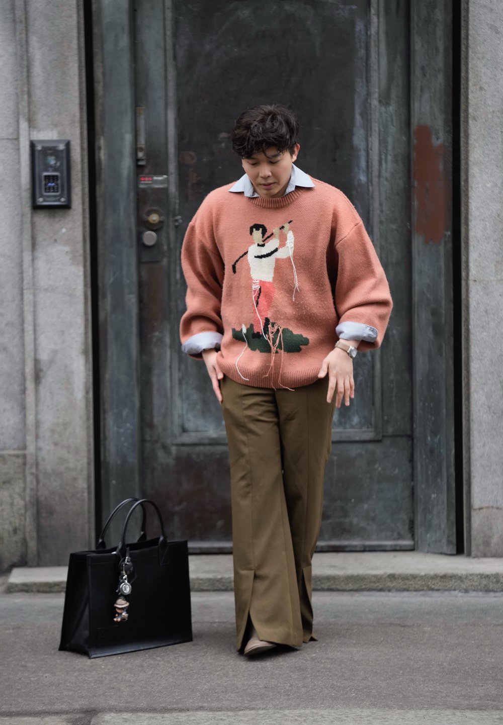 Persiku krāsas adīta džemperis ar izšūtu figūru, pelēka apkakles krekls, brūni bikses un melna lielā somiņa ar piekariņu detaļu.