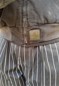 Črne delovne hlače iz tkanine s belimi črtami, ki imajo zadrgo spredaj, rebrast pas in nalepko s Carhartt logotipom zadaj.