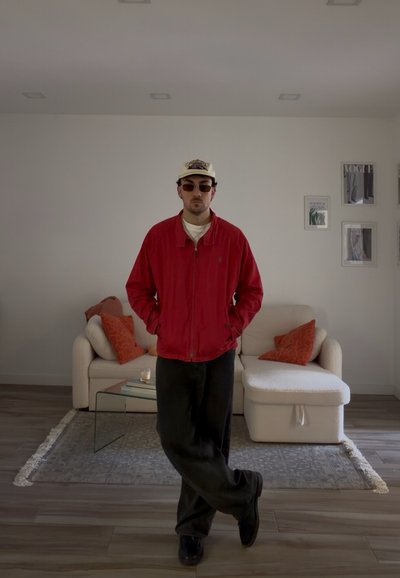 Hombre con chaqueta roja, gafas de sol y gorra, de pie con las piernas cruzadas en una sala de estar moderna con sofá blanco y cojines naranjas.