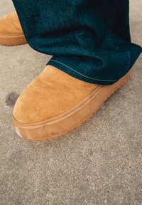 Hnedé semišové slip-on topánky s okrúhlym špicom, štruktúrovaným zvrškom a gumovou podrážkou, spárované s tmavomodrými džínsami, ktoré čiastočne zakrývajú topánku.