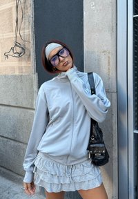 Nuori henkilö, jolla on ylisuuressa koossa olevat lasit, harmaa hiuspanta, vaaleanharmaa vetoketjullinen takki ja röyhelöhame, seisoo kaupungin rakennuksen seinää vasten.