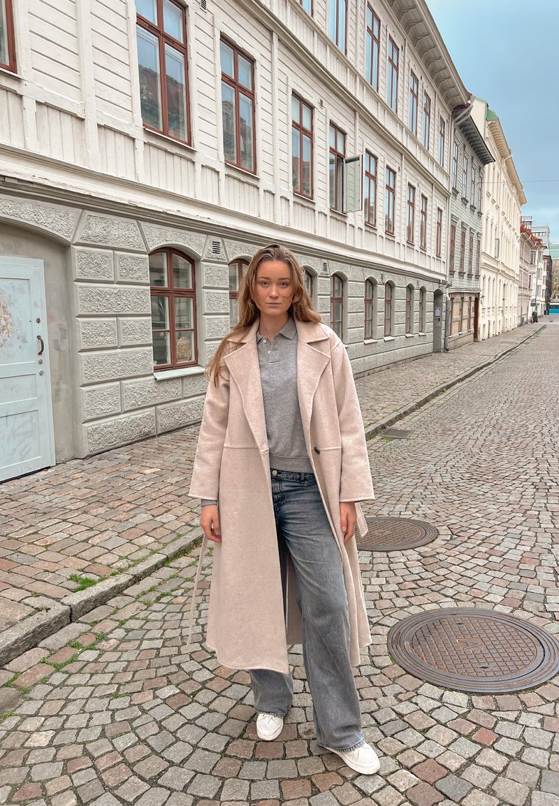 Ung kvinde iført en beige lang frakke, grå skjorte og jeans, stående på en brostensbelagt gade omgivet af hvide bygninger med vinduer.