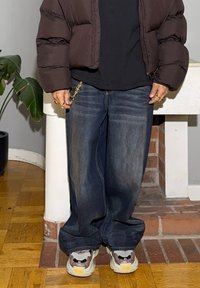 Tumma toppatakki, musta paita, haalistuneet siniset leveälahkeiset farkut ketjuyksityiskohdilla ja paksupohjaiset lenkkarit, joissa on maastokuvio ja monivärinen pohja.