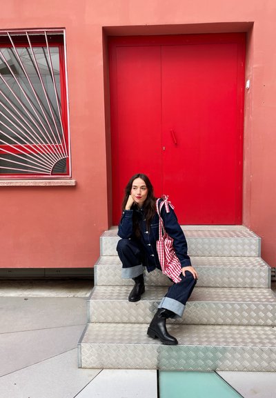 Conjunto de denim con puños enrollados, botas de cuero negro y un bolso a rayas rojas y blancas. El fondo incluye una puerta roja y escalones con un patrón.