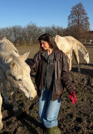 Una mujer con chaqueta de cuero marrón y vaqueros toca suavemente un caballo blanco en un campo embarrado bajo un cielo azul claro. Lleva guantes rojos.