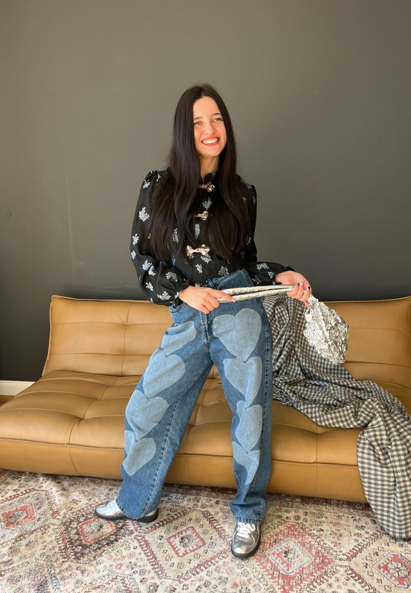 Hochgeschnittene blaue Jeans mit hellblauen Herz-Patches, kombiniert mit einem schwarzen Shirt mit floralen Stickereien. Metallic-silberne Handtasche und Schuhe.