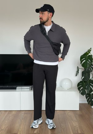 Hombre con barba que lleva una gorra negra, suéter gris, bolso cruzado negro, pantalones negros y zapatillas blancas, de pie en interiores cerca de un televisor y una planta.