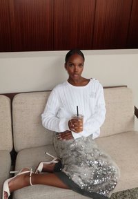 Vit kabelstickad tröja med knappar på axlarna, grå paljettkjol och vita klackar med remmar. Håller en genomskinlig kopp med ett svart sugrör.