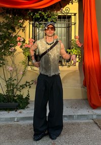 Model wearing a grey sleeveless top and wide black trousers, accessorised with multiple silver chains and bracelets, set against a floral backdrop.