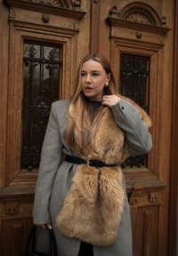 Femme aux longs cheveux blonds portant un manteau gris ceinturé d'une ceinture noire et un grand manteau en fourrure beige, debout devant une porte en bois ornée.