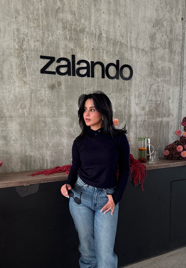 Une femme en haut bleu marine et en jeans tient des lunettes de soleil sous une enseigne Zalando. Derrière elle, des fleurs et une boisson dans un verre se trouvent sur le comptoir.