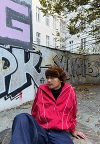 Červená bunda na zip s bílými pruhy, černý rolák a hnědá kožešinová čepice; sedí před graffiti na texturované zdi.