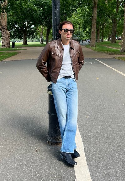 Chaqueta de cuero marrón, camiseta blanca, jeans de campana azul claro y botines negros. De pie junto a un farol en un ambiente de parque.