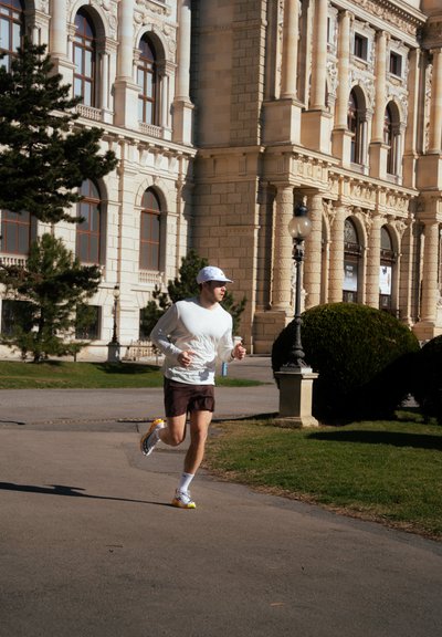 Un corredor masculino con una camiseta de manga larga blanca y pantalones cortos marrones, que lleva zapatillas con detalles en naranja, corre por un sendero cerca de un edificio clásico.