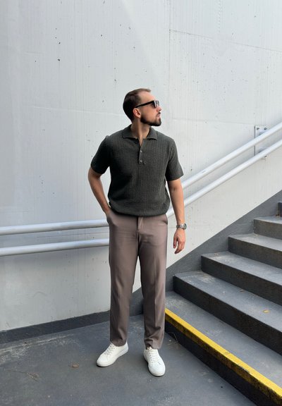 Polo de punto verde oscuro con mangas cortas, combinado con pantalones marrón claro y zapatillas blancas, de pie en unos escalones grises con borde amarillo.