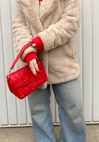 Κόκκινη δερμάτινη τσάντα με ανάγλυφο λογότυπο, ορθογώνιο σχήμα, κοντός ιμάντας, κρατημένη στο χέρι. Φοριέται με μπεζ ψεύτικο γούνινο παλτό και κόκκινες μανσέτες.