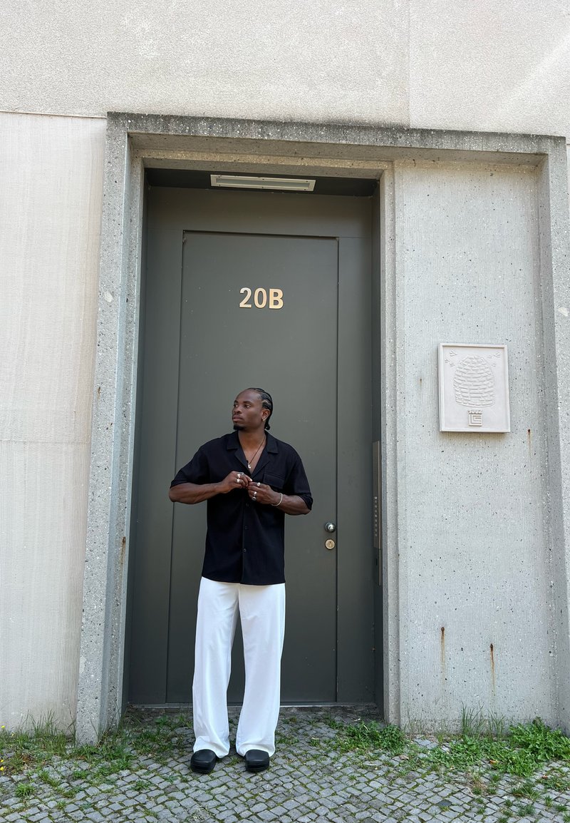Μαύρο κοντομάνικο πουκάμισο, λευκά παντελόνια με φαρδιά γραμμή και σκούρα παπούτσια, στέκεται δίπλα σε μια γκρίζα πόρτα με την ένδειξη "20B" και έναν υφασμένο τοίχο δίπλα της.