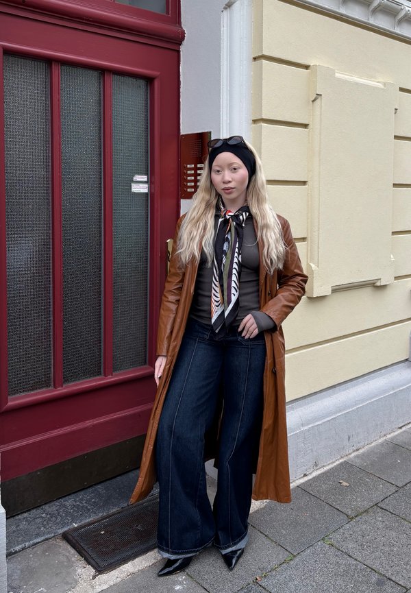 Brauner Leder-Trenchcoat, graues Oberteil, schwarze weit geschnittene Jeans, schwarzer Schal mit Feder-Muster und spitze schwarze Absätze. Vor einer roten Tür.