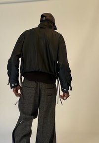 Persoană întoarsă, purtând o jachetă din piele neagră, o pălărie maro și pantaloni gri largi cu tăieturi aproape de glezne, pe un fundal simplu.