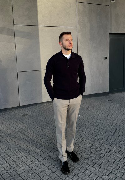 Hombre con camisa negra de manga larga, pantalones gris claro y zapatos negros, de pie con las manos en los bolsillos sobre pavimento de baldosas junto a una pared gris.