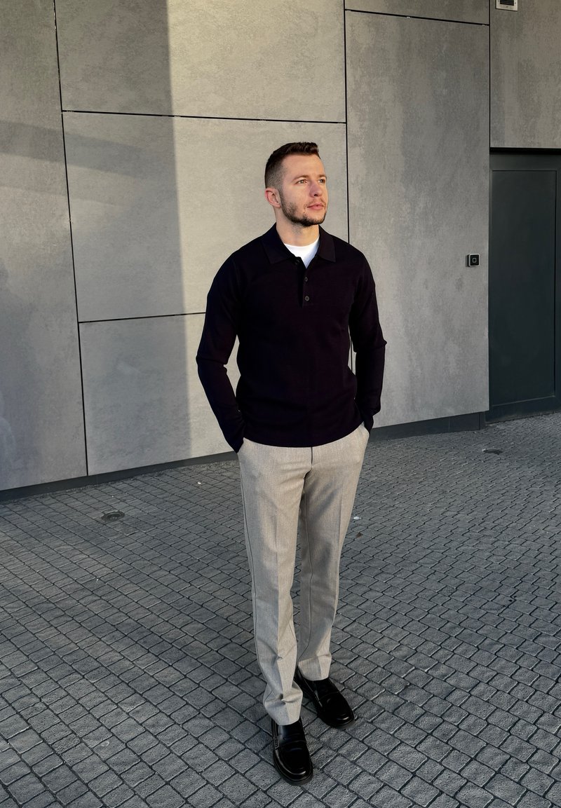 Man wearing black long-sleeve shirt, light grey trousers, and black shoes stands with hands in pockets on tiled pavement by grey wall.