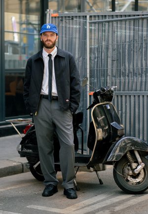 Chaqueta negra sobre una camisa blanca, corbata negra, pantalones grises y zapatos negros. Un gorro azul y una scooter negra con acentos cromados en el fondo.