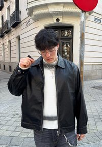 Veste en cuir noir avec un col, portée sur un pull blanc texturé. L'individu porte des lunettes et plusieurs colliers en superposition.