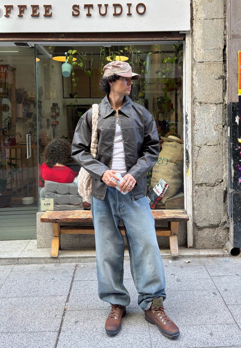 Jeune personne portant une veste en cuir et un jean large, debout à l'extérieur d'un café, tenant une tasse et regardant sur le côté.
