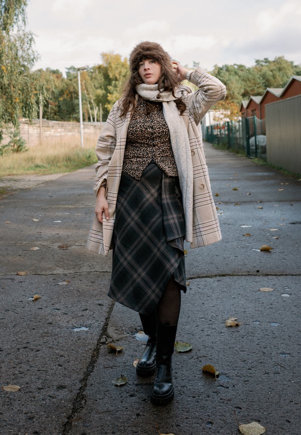 Une femme portant un bonnet de fourrure, une écharpe, un manteau à carreaux et une jupe se tient sur un sentier jonché de feuilles, à côté d’arbres d’automne et de garages.