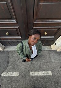 Giacca bomber verde oliva, camicia bianca, pantaloni neri; in piedi su una strada grigia; porte in legno scuro con maniglie in ottone sullo sfondo.