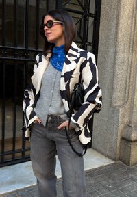 Femme portant un blazer à motifs noirs et blancs, un pull gris, un bandana bleu, des lunettes de soleil foncées et un jean gris, debout près d'une grille métallique.