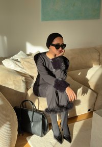 Ensemble tricot noir avec accents en fourrure aux poignets et à l'ourlet. Talons aiguilles noirs élégants, lunettes de soleil oversized et sac à main noir. Fond de canapé ton neutre.