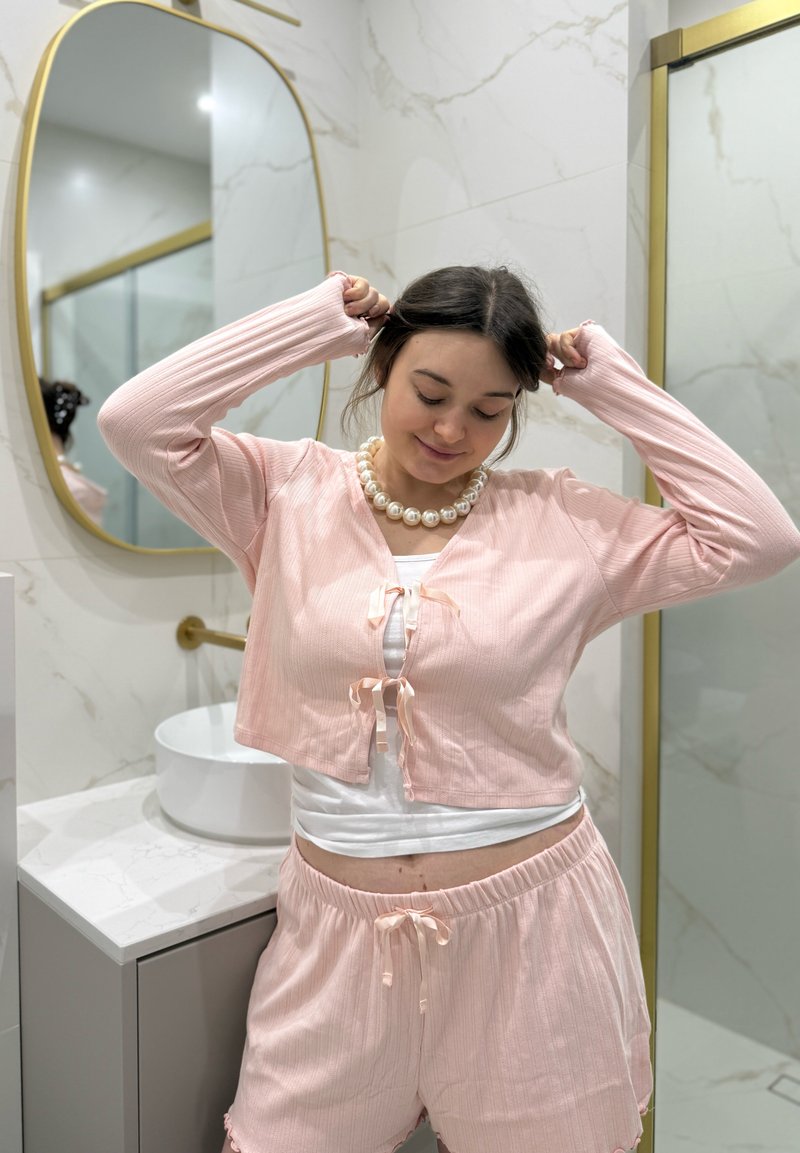 Cardigan côtelé rose avec liens à l'avant, assorti à un short. Tissu lisse, coupe ample, porté sur un haut blanc, dans un cadre de salle de bains.