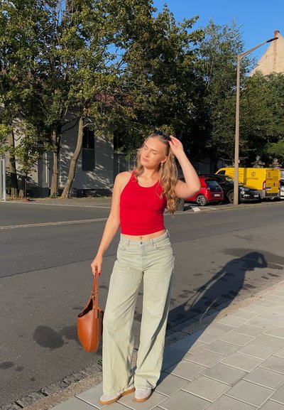 Top rojo sin mangas, jeans de pierna ancha verde claro y zapatillas beige. Bolsa de mano marrón. El fondo presenta árboles y coches aparcados en una calle.