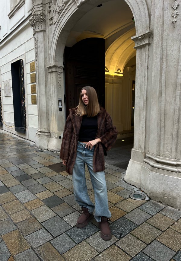 Žena v hnědém kožichu, černém topu, světle modrých džínách a hnědých botách stojí na dlážděné ulici u zdobeného kamenného obloukového vchodu.