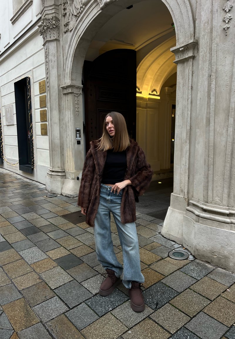 Ženska, oblečena v rjav krznen plašč, črno majico, svetlo modre kavbojke in rjave škornje, stoji na tlakovani ulici ob okrašenem vhodu iz kamnitega loka.