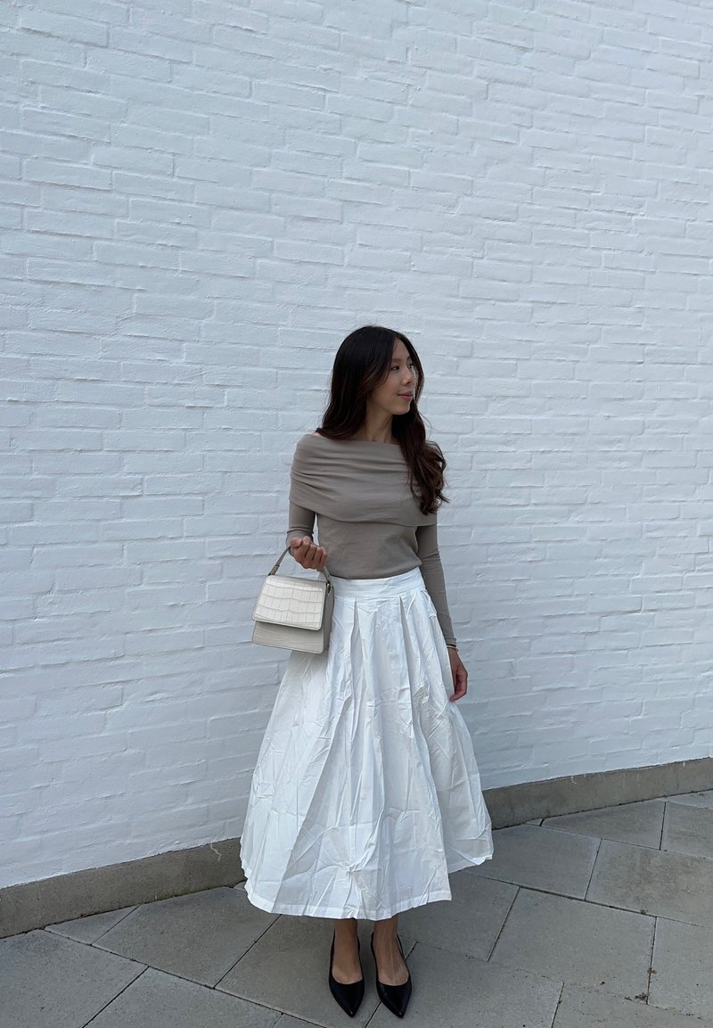 Μπεζ μπλούζα με μακριά μανίκια, λευκή πλισέ φούστα με γυαλιστερή επιφάνεια, μαύρες γόβες με τετράγωνη μύτη και μικρή λευκή τσάντα με υφασμάτινη υφή.