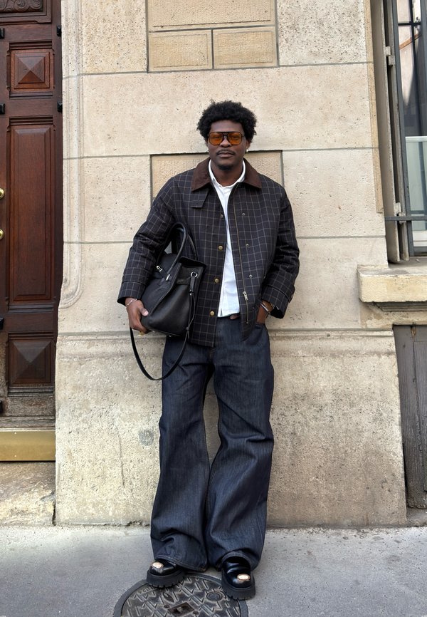 Mann mit Sonnenbrille, kariertem Jackett, weissem Hemd, weiter Jeans und schwarzen Schuhen steht an einer beigen Steinmauer und hält eine schwarze Handtasche.