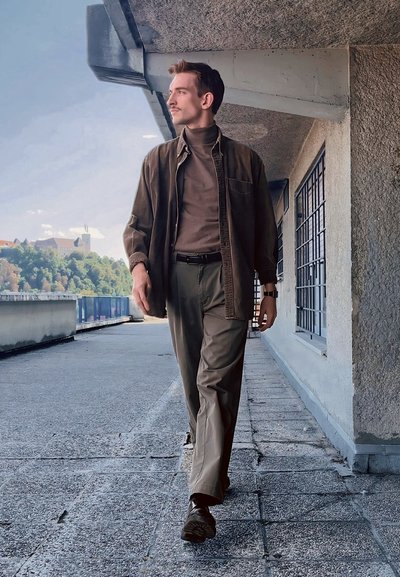 Hombre caminando por un camino de concreto, vistiendo una camisa marrón de botones sobre un cuello alto marrón, pantalones verdes y zapatos oscuros.