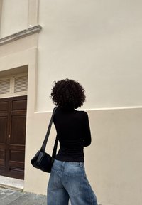 Person med krøllete hår, svart genser med lange ermer, blå jeans og svart skulderveske står vendt mot en beige bygningsvegg nær en tredør.