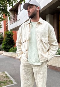 Ljust beige linnejacka med två framfickor över en ljusgrön strukturerad skjorta och matchande byxor. Outfitten kompletteras av en grå keps.