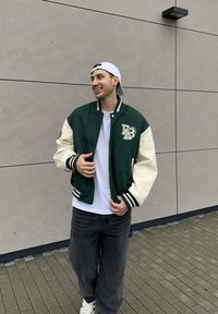 Vihreä ja kermanvärinen varsity-takki resorilla varustetuilla hihoilla, kirjotulla logolla ja napillaisella suljennalla. Päällä valkoinen t-paita ja mustat farkut.