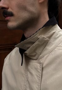 Veste beige à col montant, avec une doublure foncée et texturée. Détails notables : un rabat diagonal et un design épuré.