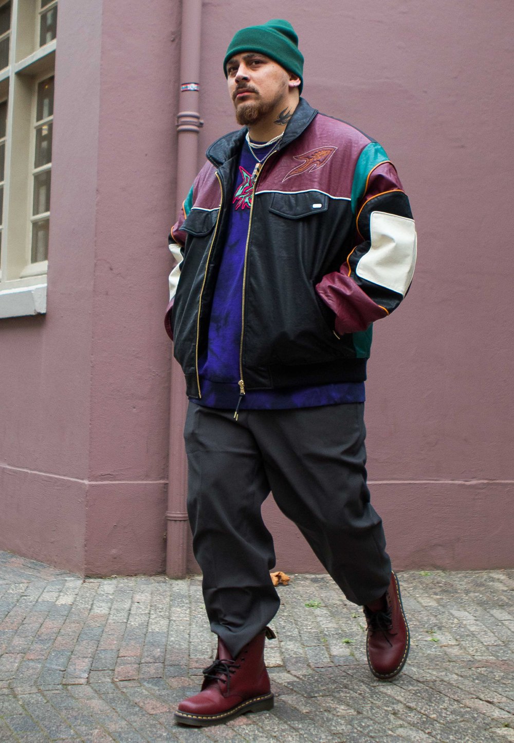Jaqueta de couro colorida com um design em bordô, verde e preto. Combinada com uma camisa roxa de tie-dye, calças cinzas largas e botas vermelhas com atacadores.