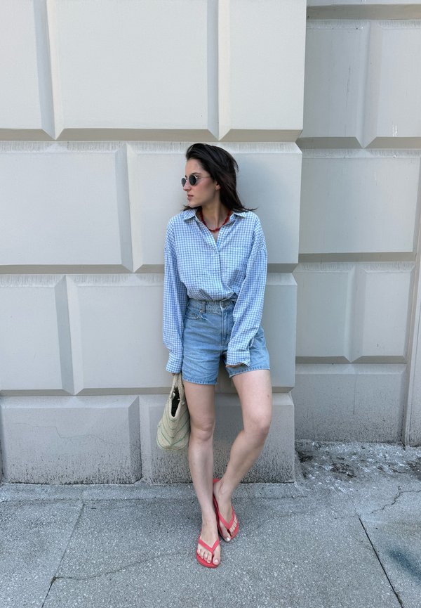 Jeune femme avec des lunettes de soleil, chemise à carreaux bleue, short en jean et tongs rouges appuyée contre un mur texturé beige tenant un sac tissé.