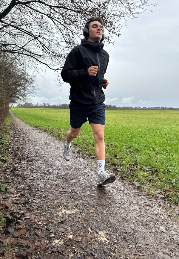 Coureur en veste imperméable noire et shorts noirs, portant des écouteurs, sur un chemin boueux à côté d'un champ gazonné avec des arbres.