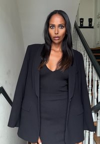 Blazer nero sopra un vestito a coste nero, con scollatura a V profonda. La modella è in piedi su una scalinata con balaustre bianche sullo sfondo.